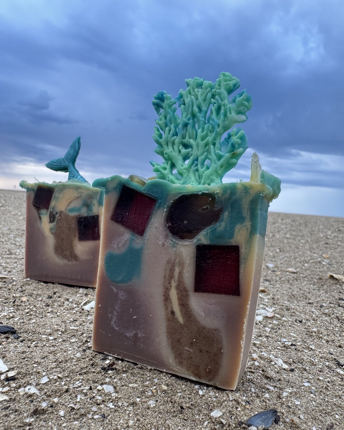Zwei handgemachte Seifen mit Koralle und Meerjungfrauenflosse stehen am Strand unter aufziehenden Regenwolken