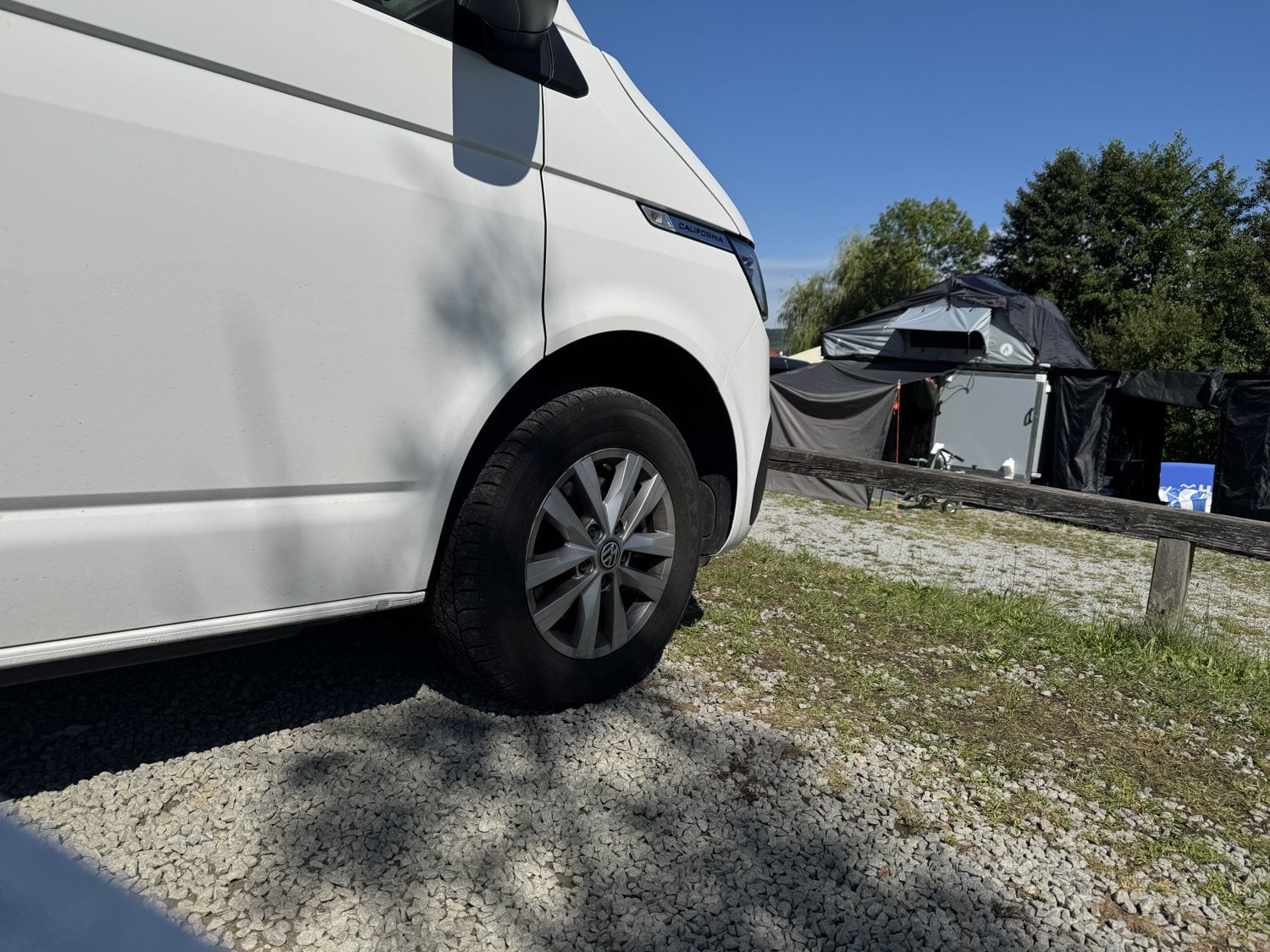 Weißer VW California Bulli auf einem Campingplatz bei blauem Himmel neben einem Zelt