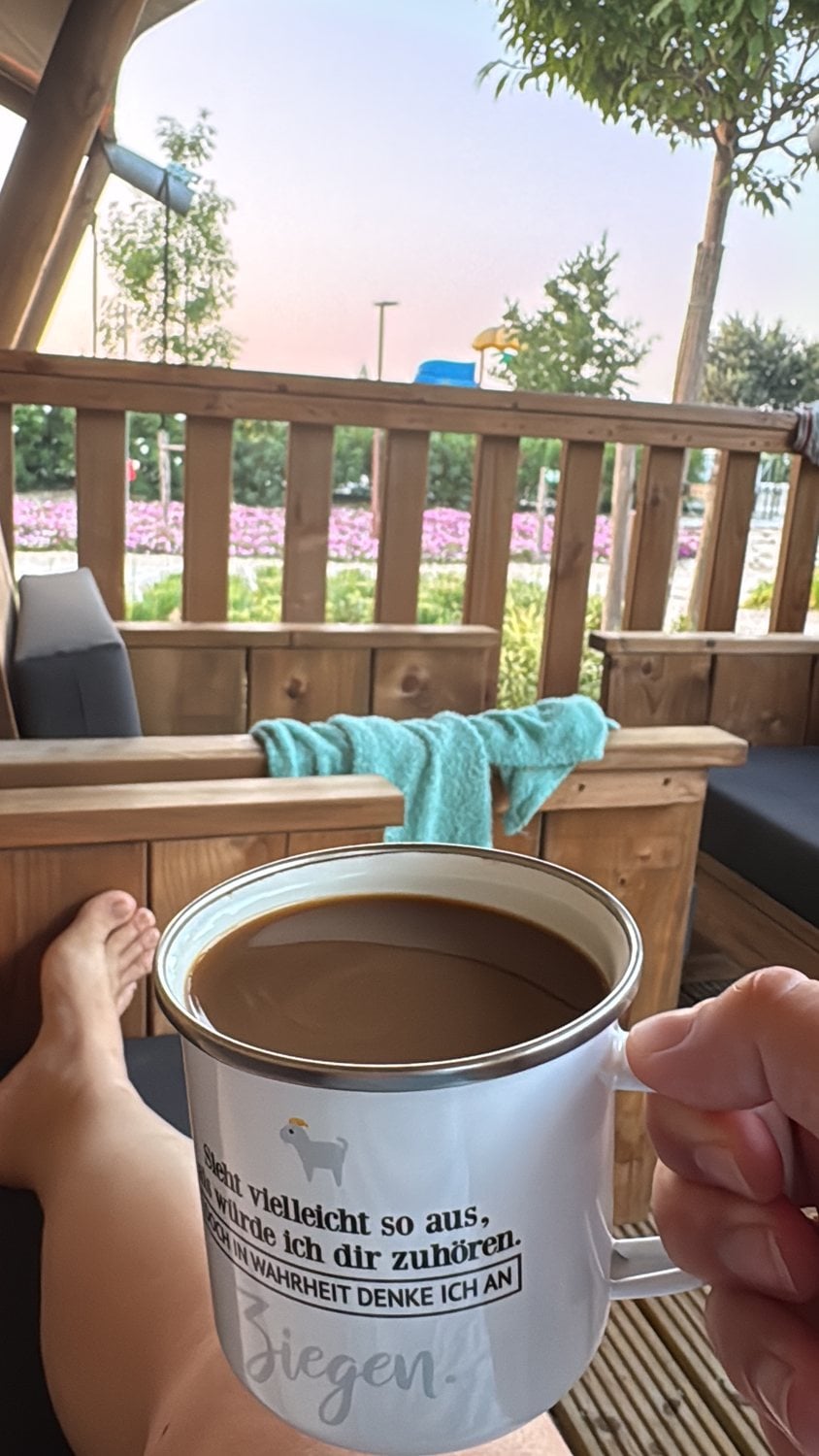 Tasse Kaffee mit Ziegen-Spruch auf einer Holzterrasse im Morgenlicht im Italienurlaub